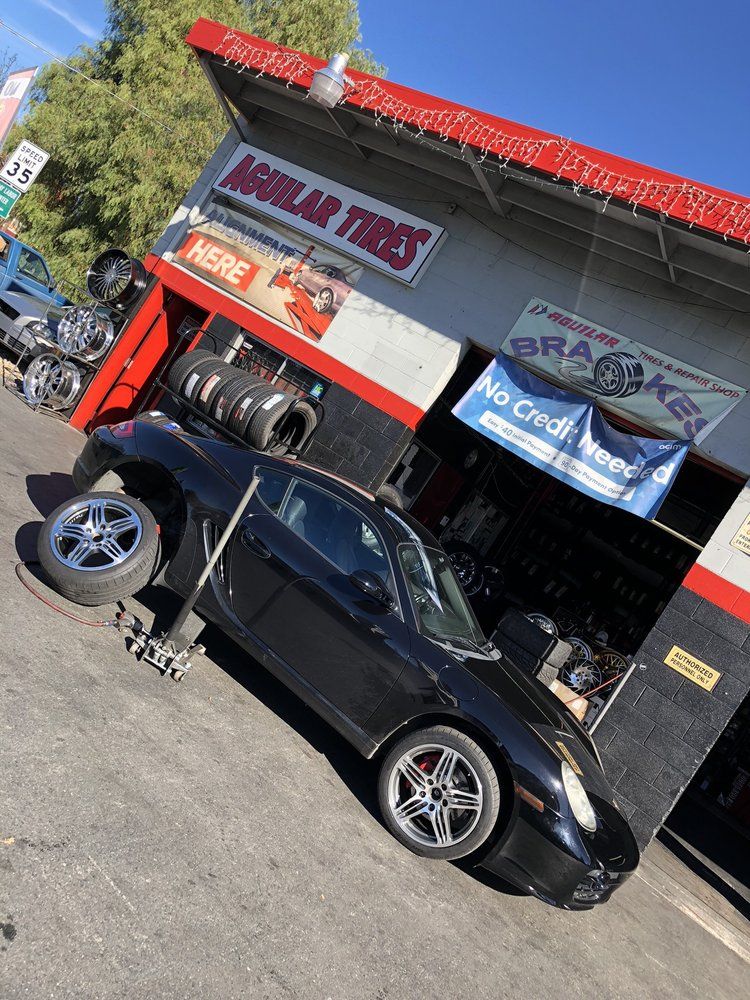 A black sports car getting tire service in front of the Aguilar Tires bays, with tires and rims stacked by the door
